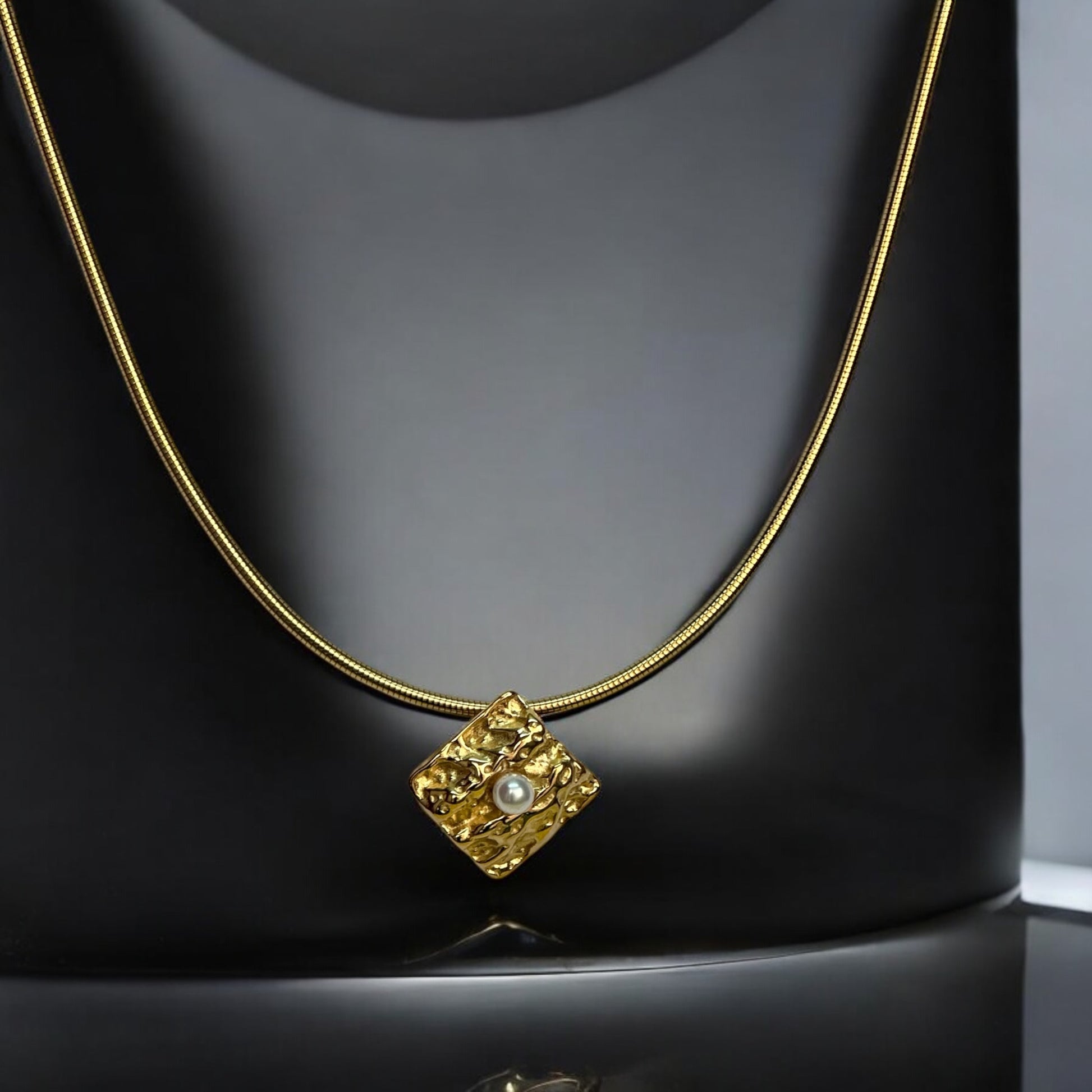 Gold-toned anti-tarnish chain necklace with a textured square pendant featuring a single pearl centerpiece, showcased on a sleek black display stand.