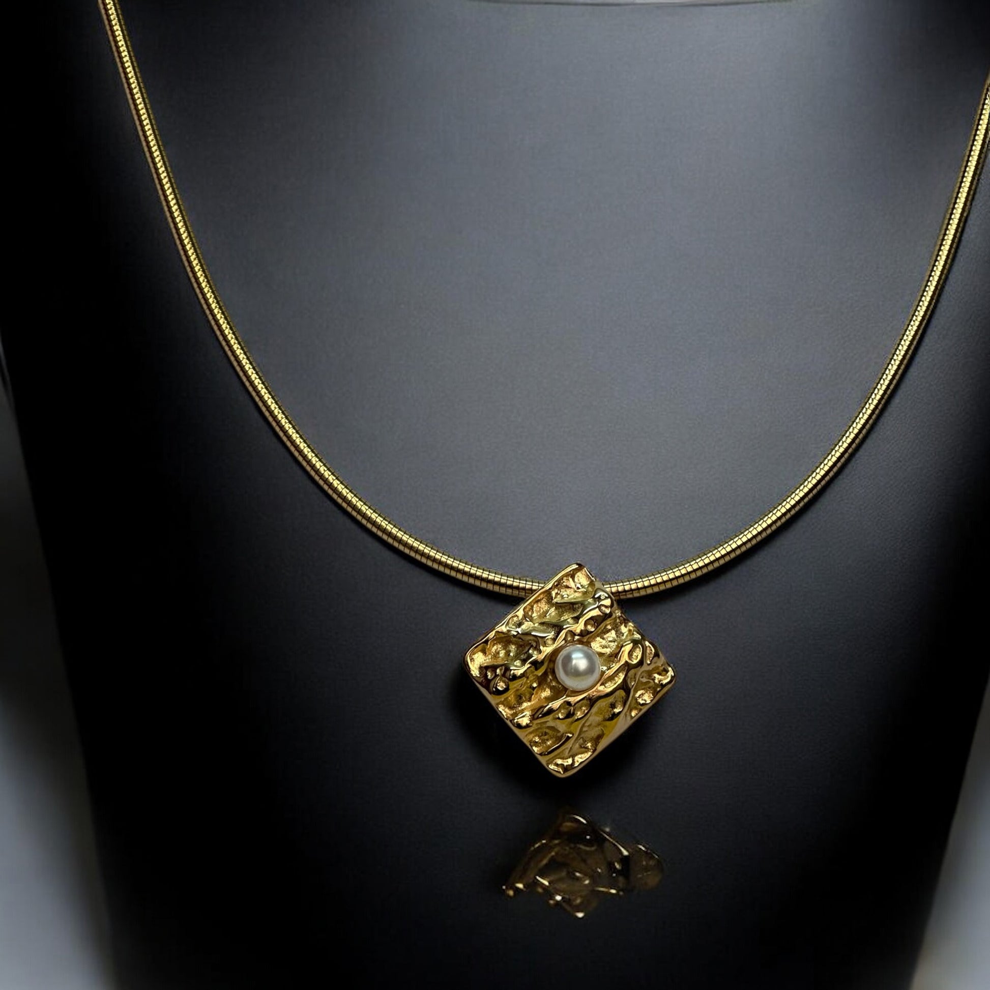 Gold-toned anti-tarnish chain necklace with a textured square pendant featuring a single pearl centerpiece, showcased on a black jewellery display stand.