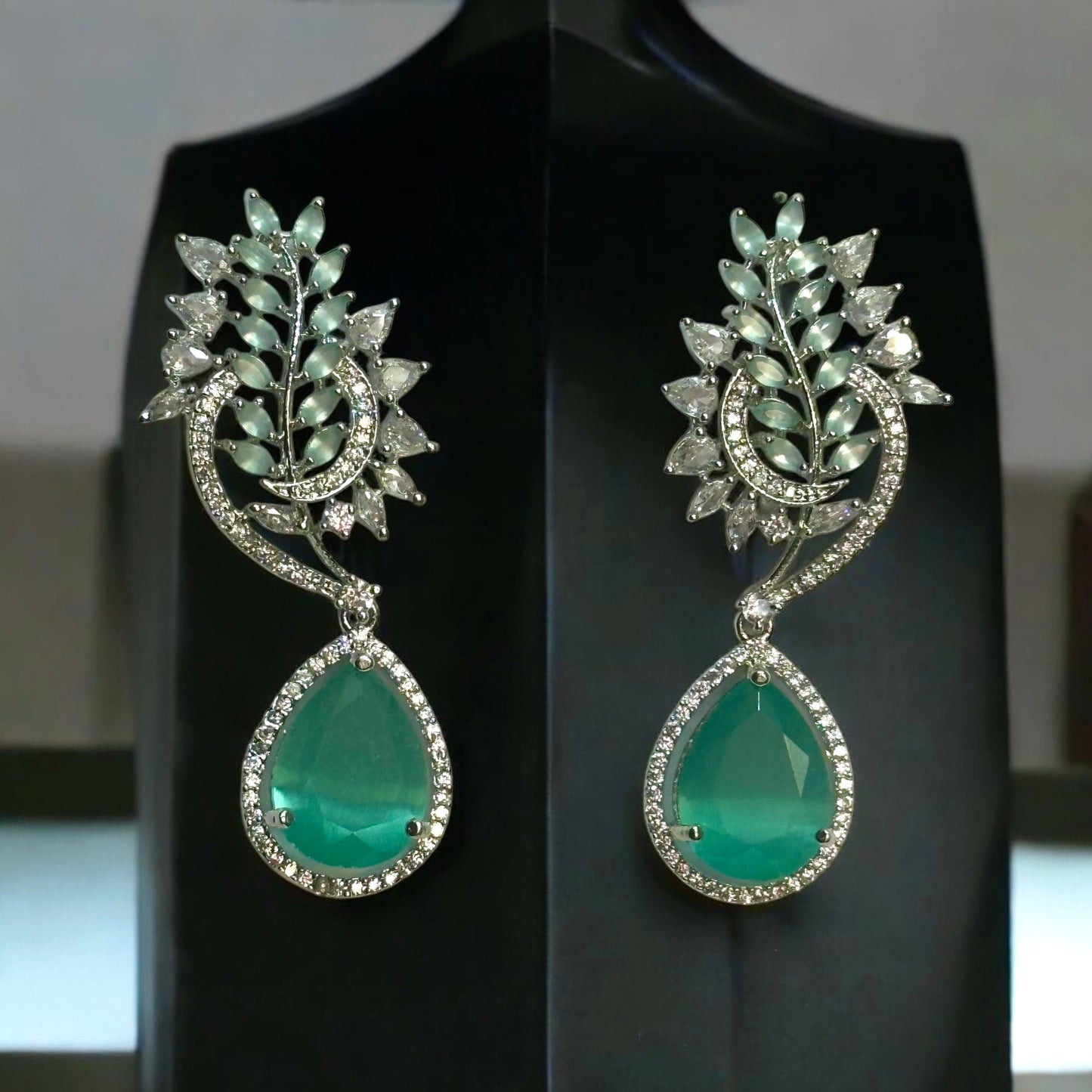 Beautiful American Diamond Statement Earrings In Greenish Magenta Shade placed on a black coloured jewellery display stand with a leaves design.