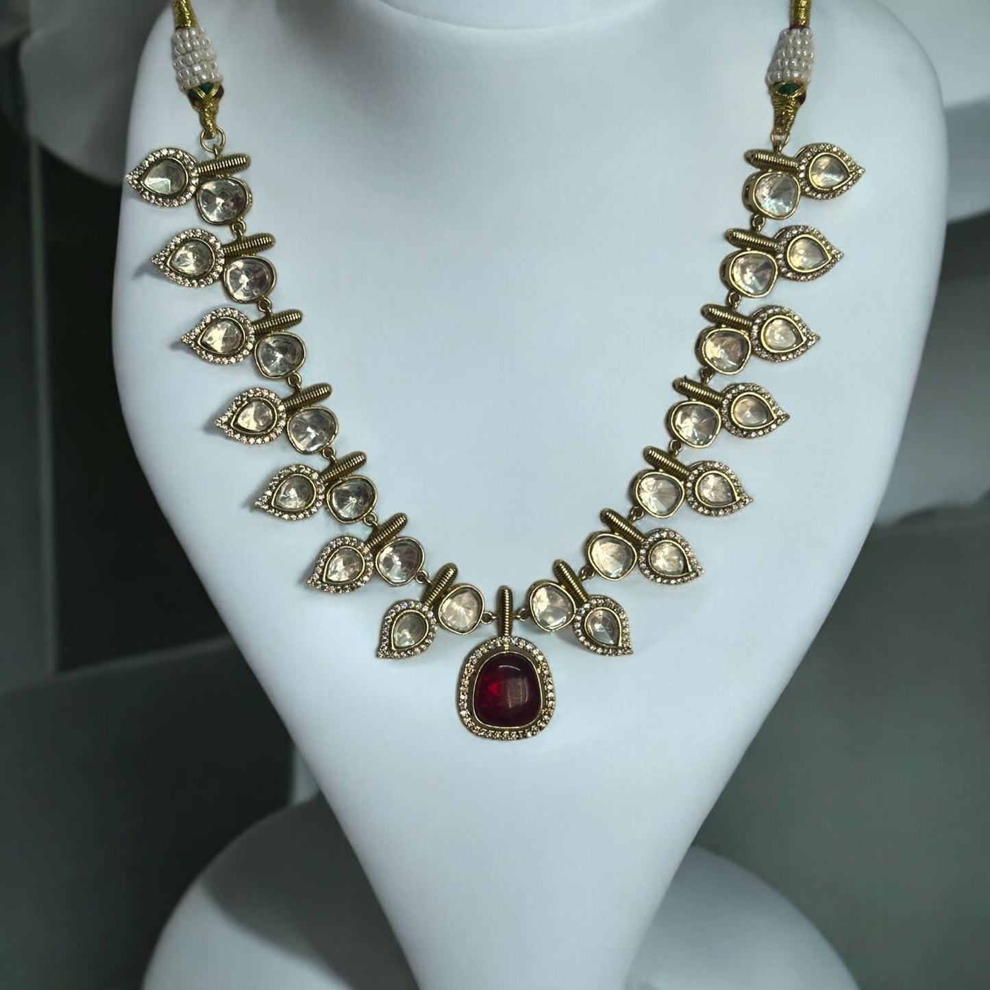 Elegant designer polki kundan necklace with teardrop accents and red gemstone centerpiece placed on a jewellery display stand.