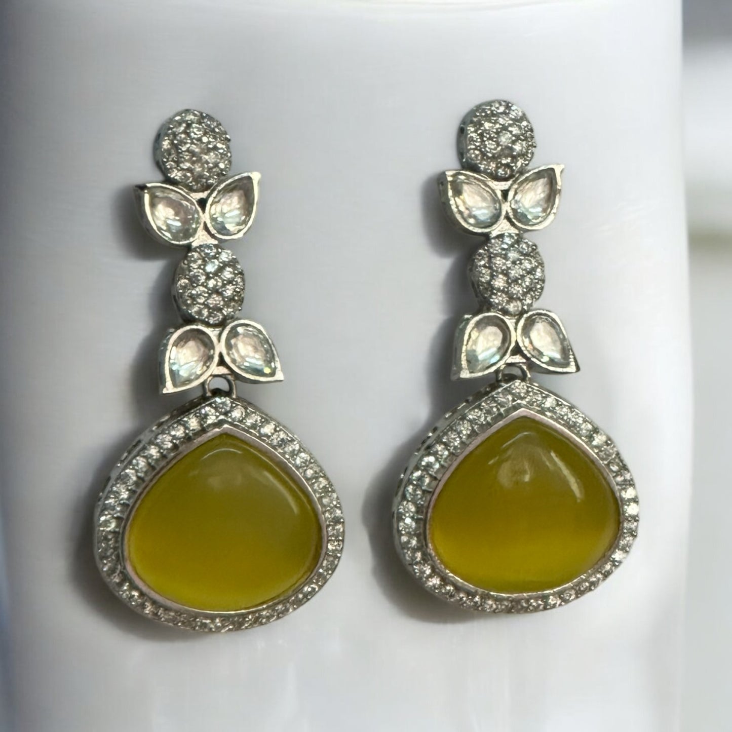 Elegant American Diamond Yellow Partywear Earrings with teardrop yellow stones and shimmering leaf-shaped studded accents on a white jewellery display stand.