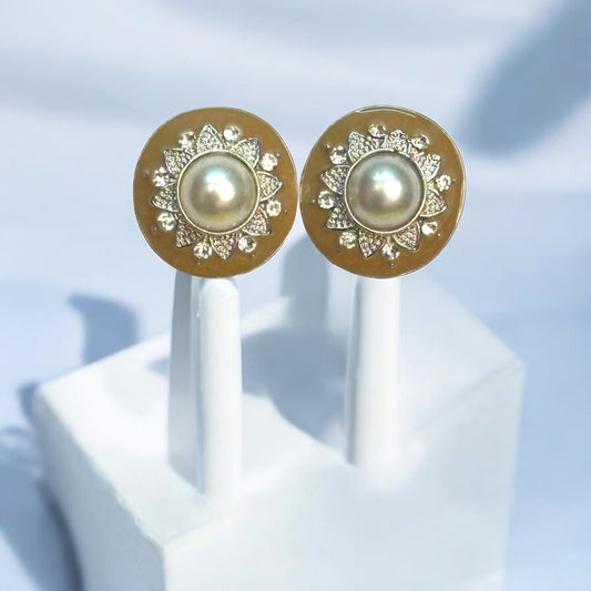 Elegant pearl stud earrings with a gold-toned circular base, featuring intricate floral detailing and embedded white stones, displayed on a white stand with a soft blue backdrop.