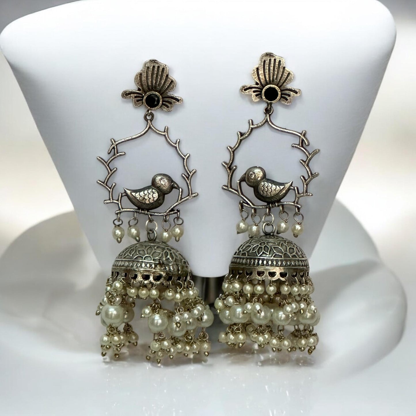 German Silver Bird Design Jhumki Earrings featuring intricate bird motifs perched on branches, adorned with dome-shaped jhumkis and cascading pearl drops, displayed on a white stand.