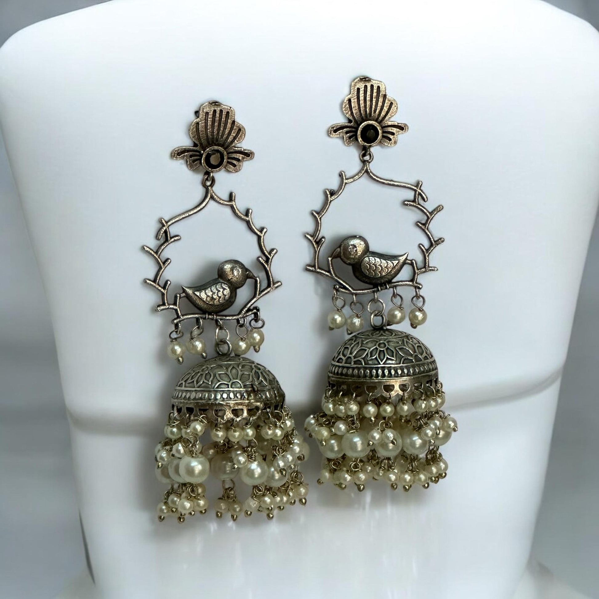 German Silver Bird Design Jhumki Earrings featuring intricate bird motifs perched on branches, adorned with dome-shaped jhumkis and cascading pearl drops, displayed on a white stand.