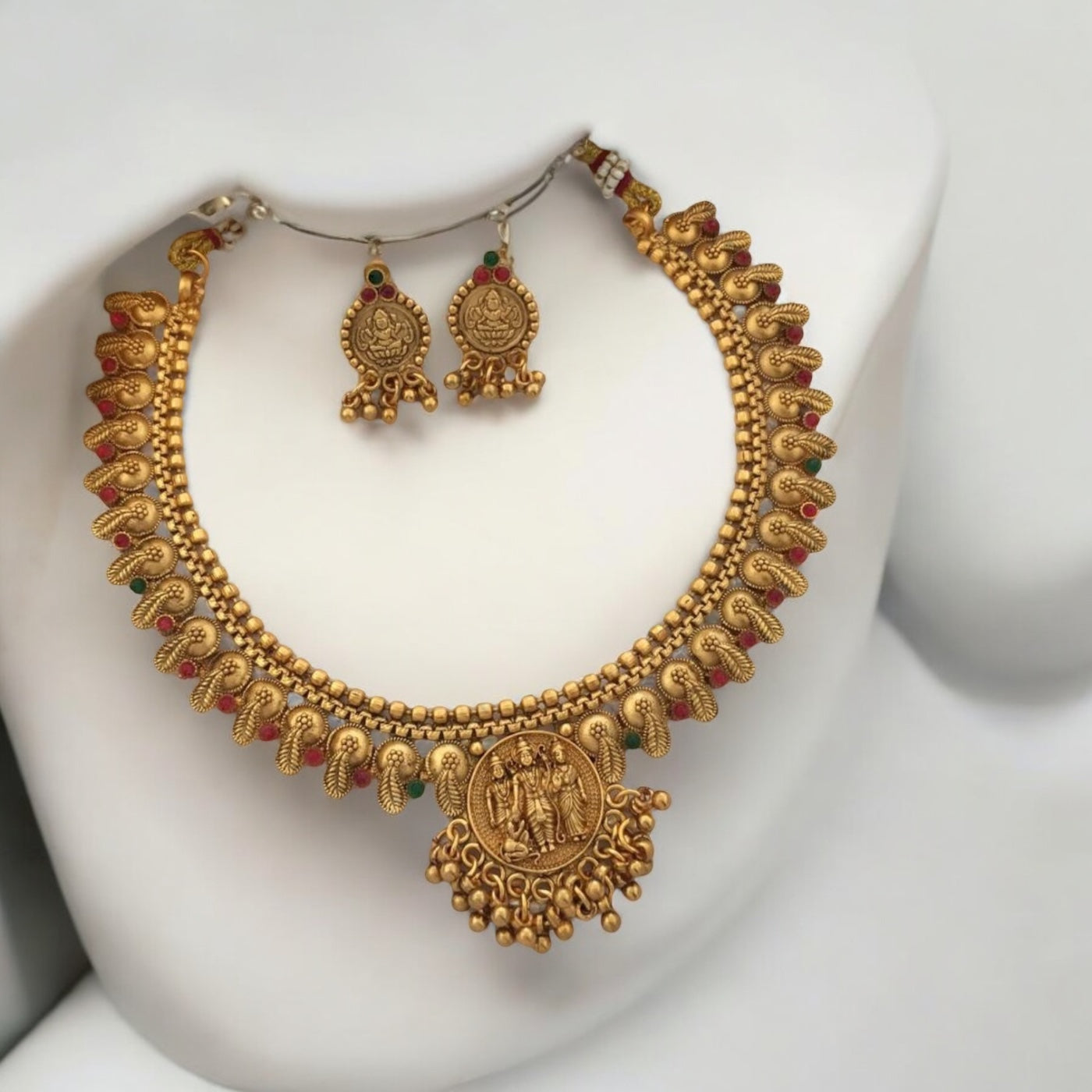 Gold-Plated Ram Darbar Temple Necklace with Earrings – traditional gold-tone necklace featuring a detailed Ram Darbar motif, paisley designs, and red-green enamel accents placed on a white jewellery display stand.. A heritage-inspired set for festive and wedding occasions.