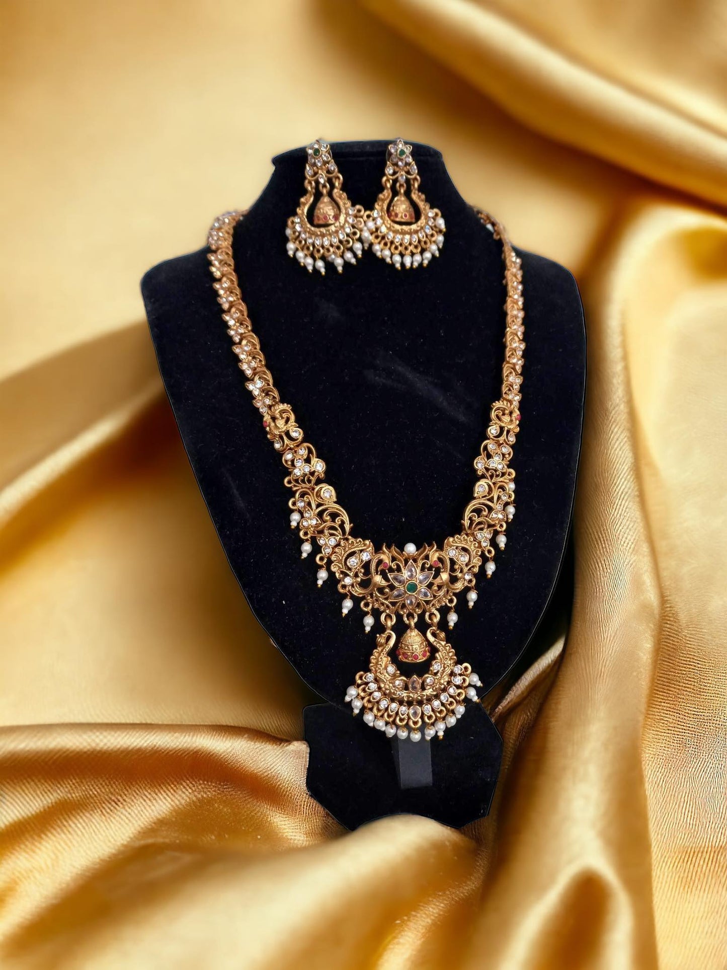 Gold-Plated Temple Necklace with Earrings – intricately designed gold-plated necklace featuring temple-inspired motifs, pearl drops, and matching earrings placed on a golden fabric background. Perfect for weddings and cultural events.