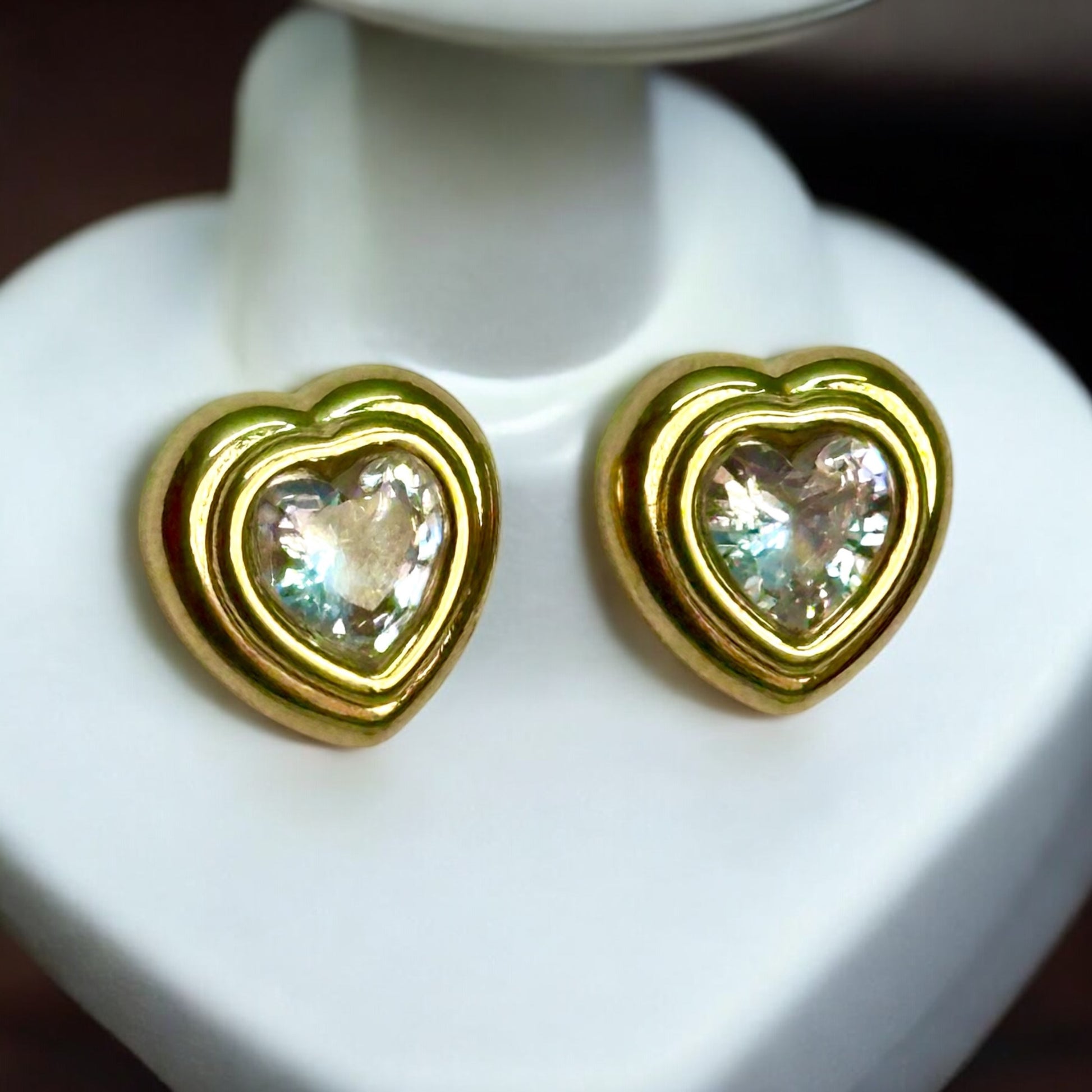 Gold plated casual wear heart-shaped earrings with an artificial shiny diamond stone in the centre. The earrings are placed on a white coloured jewellery display stand.