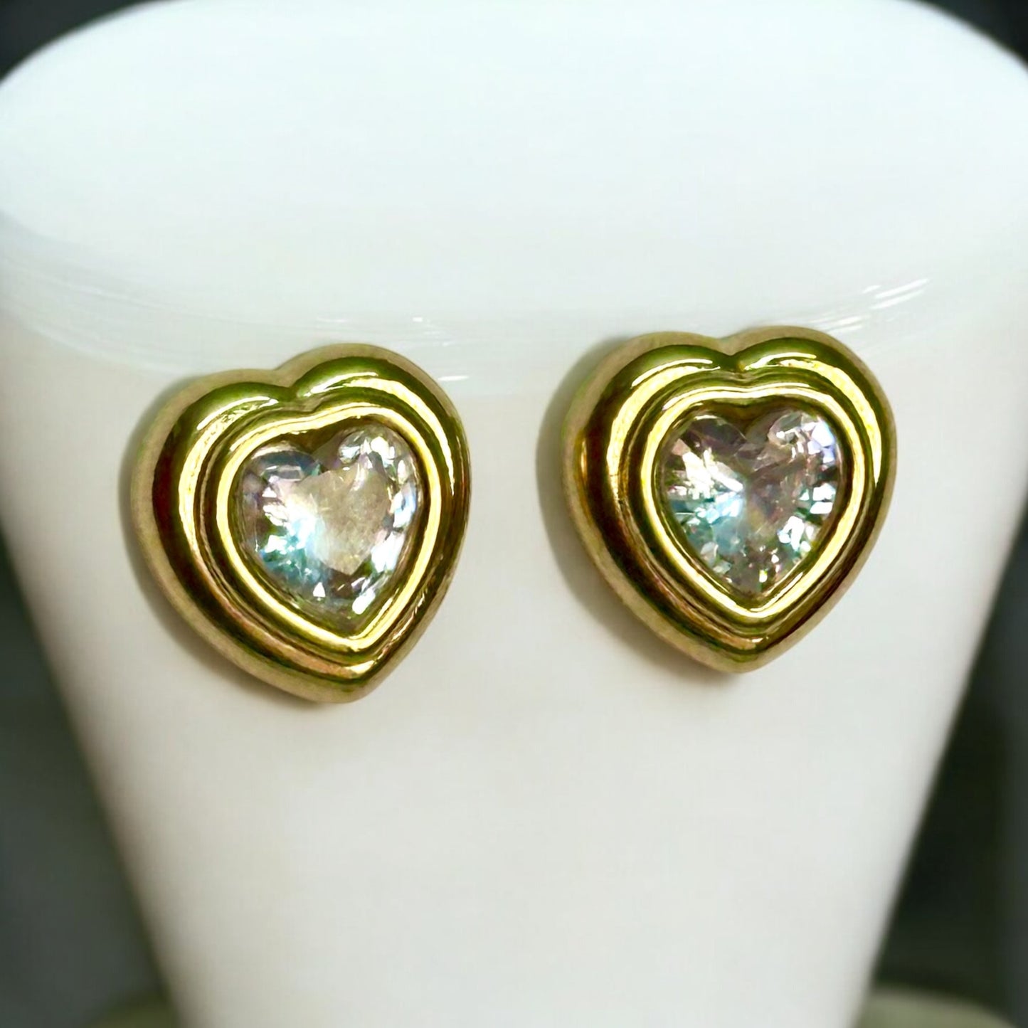 Gold plated regular wear heart-shaped earrings with an artificial shiny diamond stone in the centre. The earrings are placed on an off white coloured jewellery display stand.