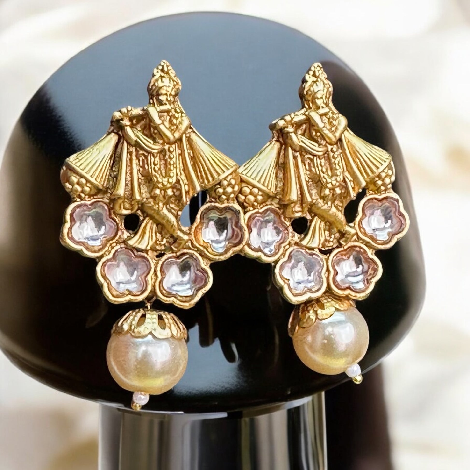 Gold Toned Earrings With Krishna Design featuring silver accents and a gold-toned design intricately depicting Lord Krishna. Displayed on a black stand, showcasing the elegant craftsmanship and divine design.