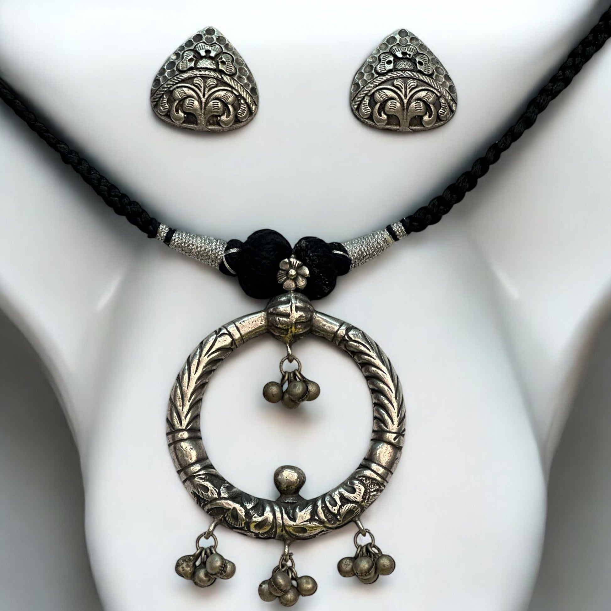 Handmade silver replica necklace featuring a bold circular pendant with intricate engravings and ghungroo charms, paired with detailed triangular stud earrings. The set is displayed on a white mannequin with a braided black thread chain.