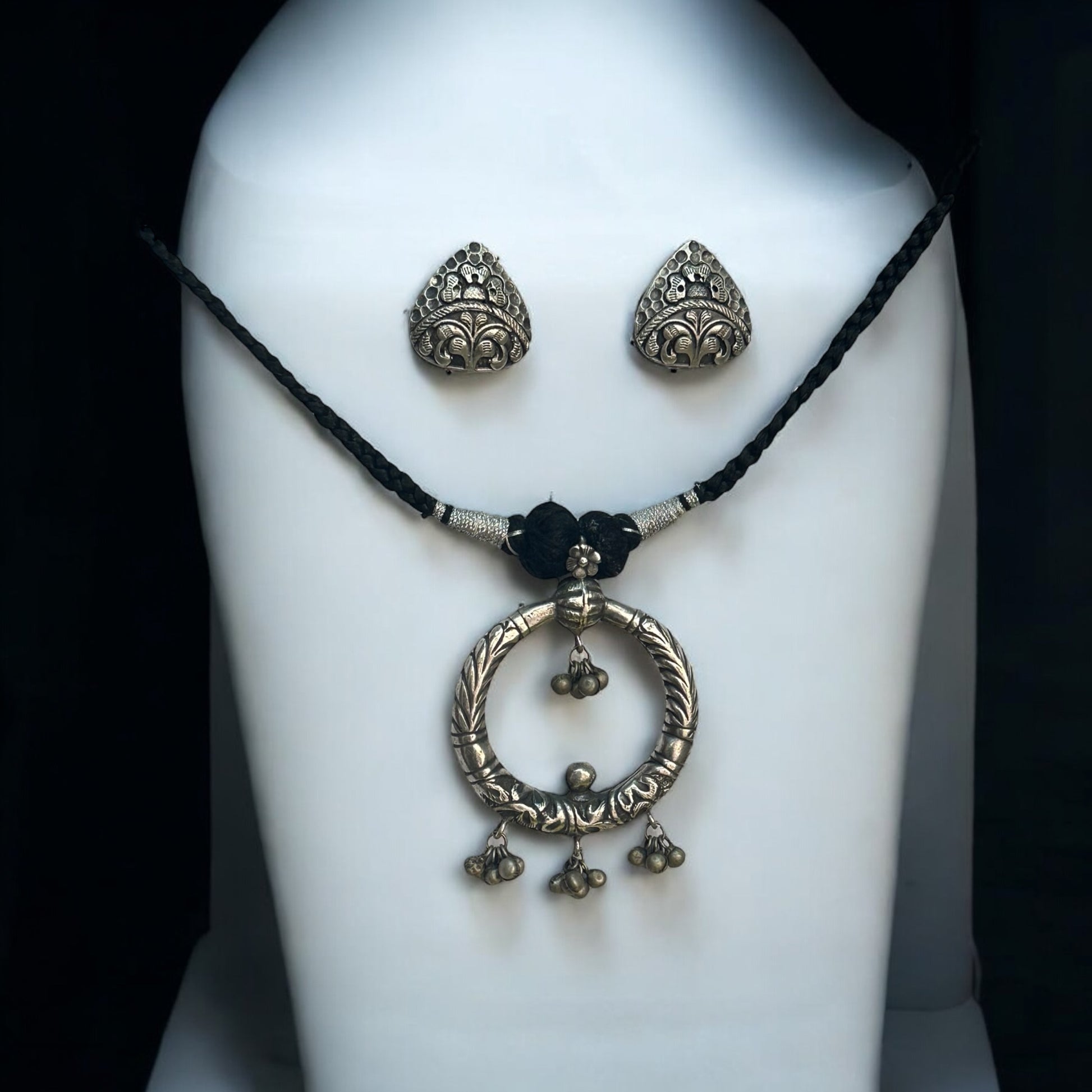 Handmade silver replica necklace featuring a bold circular pendant with intricate engravings and ghungroo charms, paired with detailed triangular stud earrings. The set is displayed on a white jewellery display stand with a braided black thread chain.