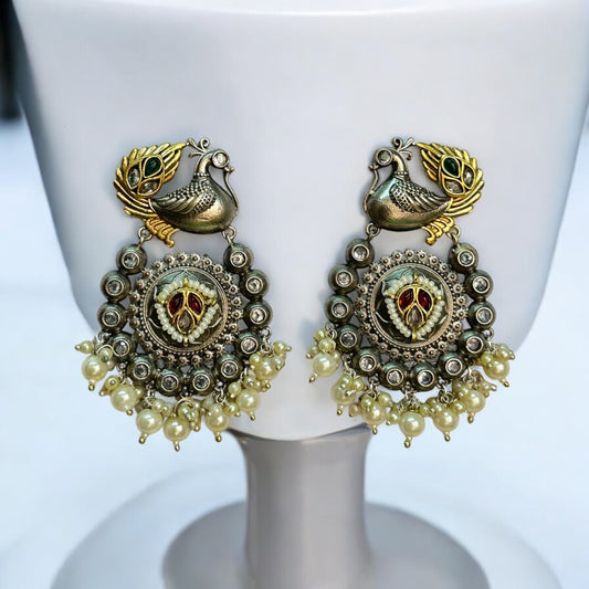 Jadau Kundan dual-tone statement earrings featuring peacock motifs, intricate circular detailing, red and green enamel work, and pearl hangings, displayed on a white jewelry stand with a soft blue background.