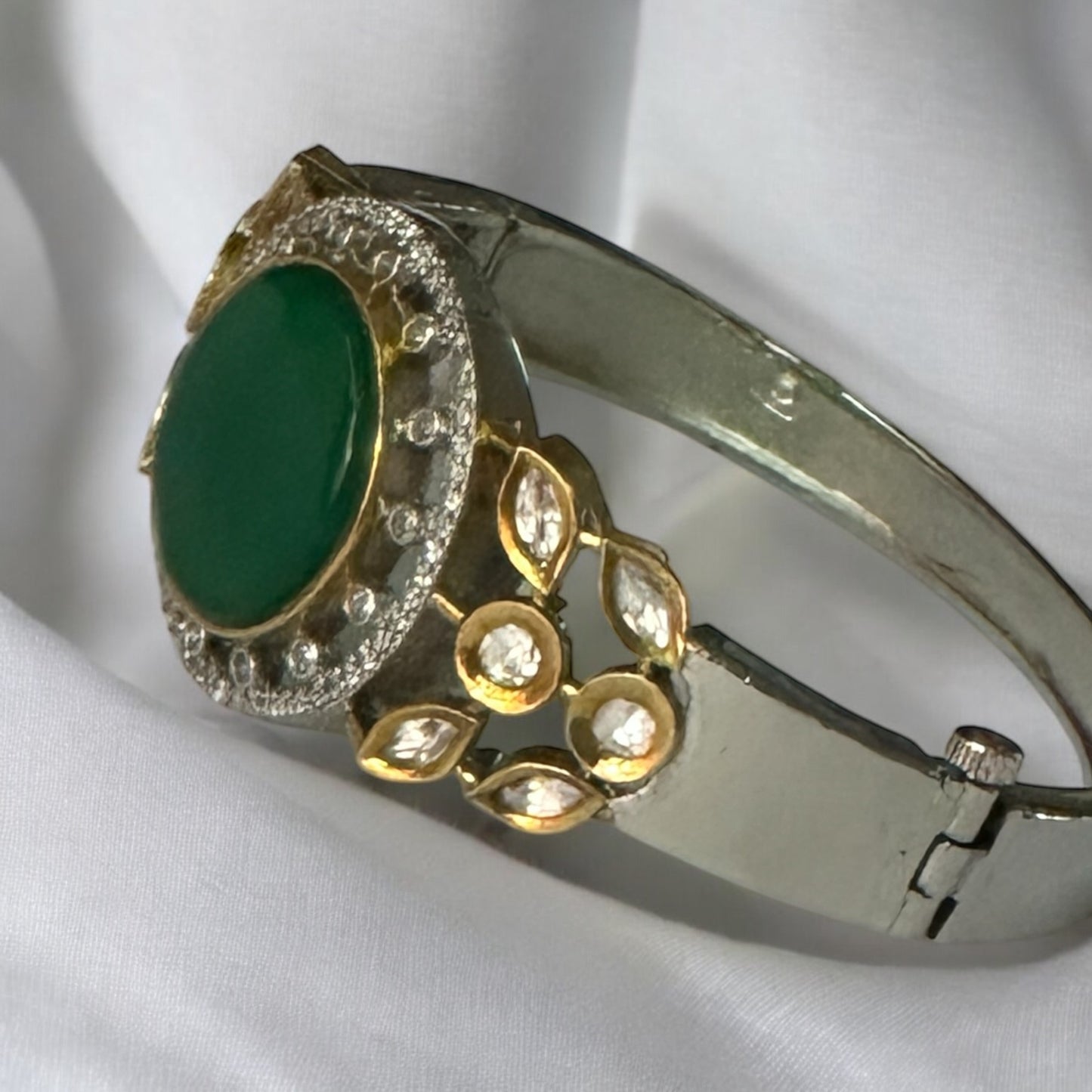 Lying on a white fabric an elegant Moissanite Bracelet featuring a vivid green Monalisa stone centerpiece with intricate gold and silver accents.