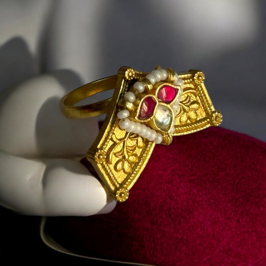 Traditional Pachi Kundan ring in gold-tone with intricate design, adorned with red and white gemstones and delicate pearl detailing. It is placed on a red velvet jewellery display stand.