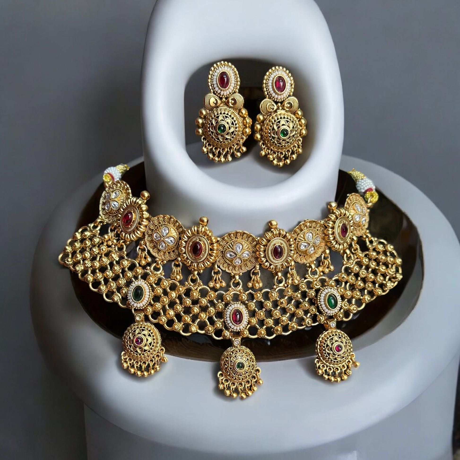 Golden Rajwadi Kundan choker necklace set with intricate detailing, red and green gemstone accents, and matching earrings. Both placed on a black and white jewellery display stand.