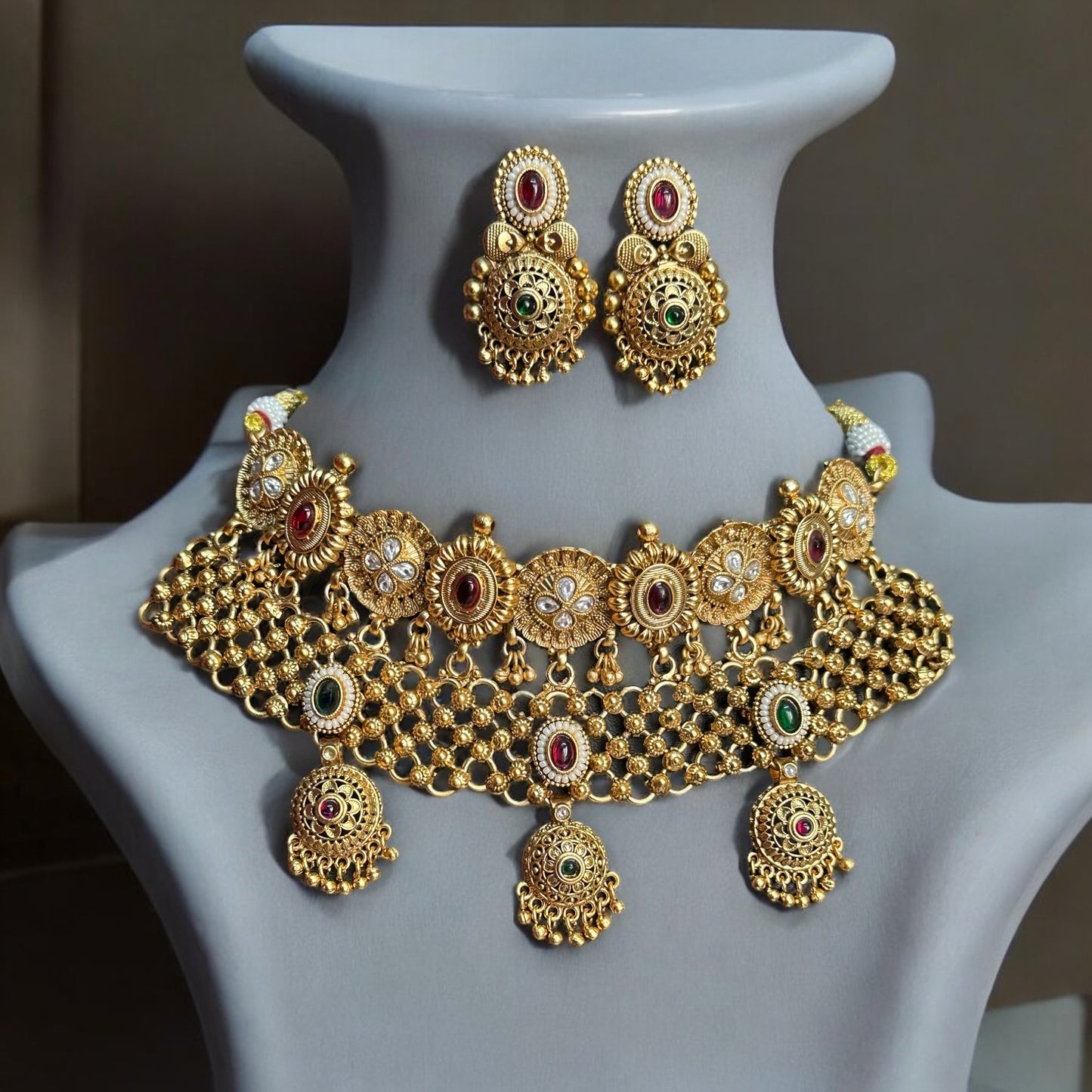 Golden Rajwadi Kundan choker necklace set with intricate detailing, red and green gemstone accents, and matching earrings. Both placed on a grey coloured jewellery display stand.