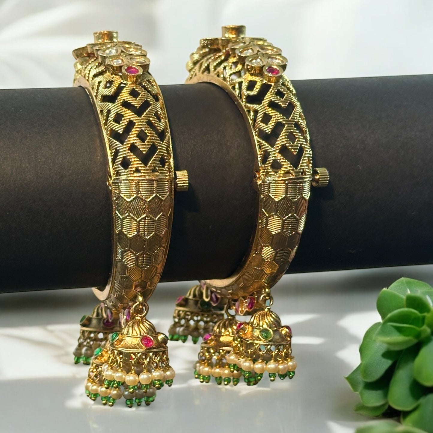 Rajwadi Premium Kundan Adjustable Bangles placed on a black roll with three jhumkas hanging from the bangle design and in background there is a white fabric and a small white plant.