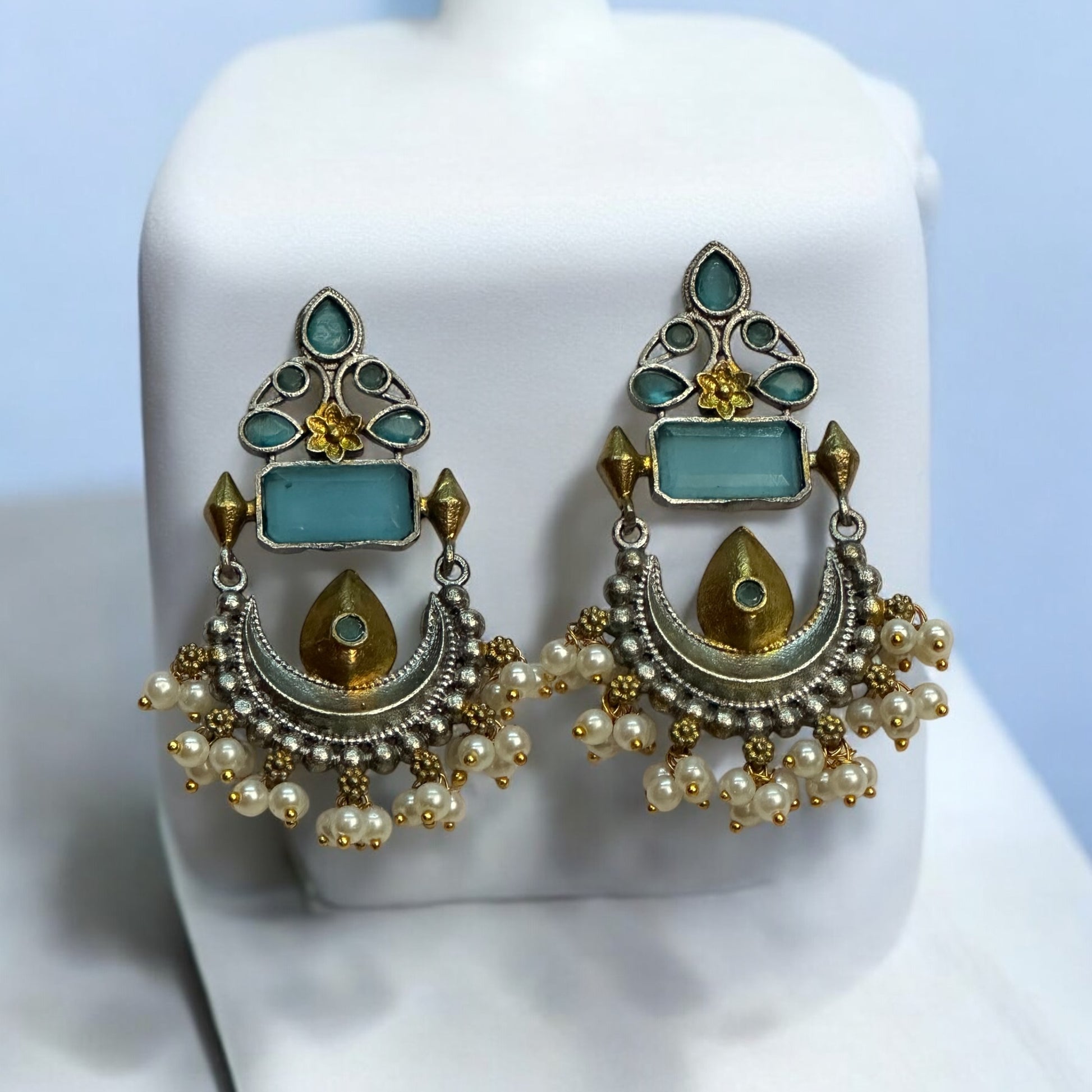 Silver Look Alike Dual Tone Statement Earrings having blue coloured stone and a gold tone with white beads placed on a white coloured jewellery display stand.