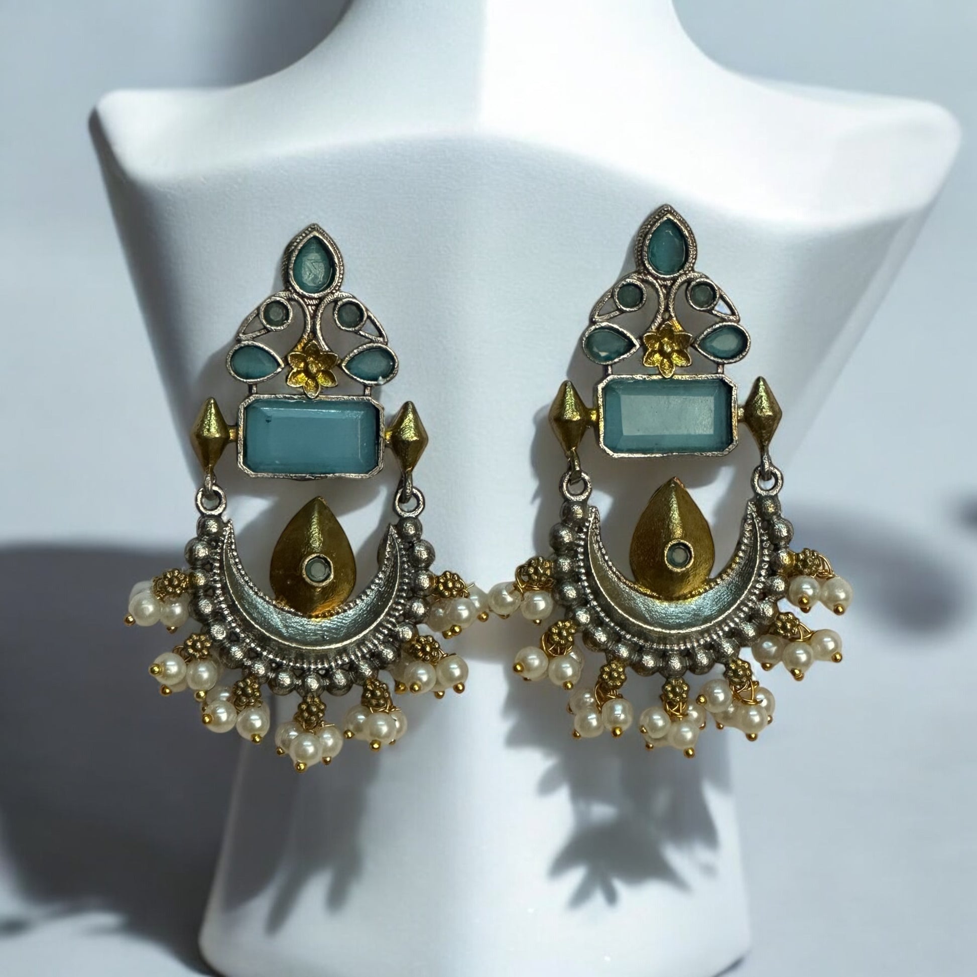 Silver Look Alike Dual Tone Statement Earrings having blue coloured stone and a gold tone with white beads placed on a white coloured jewellery display stand.