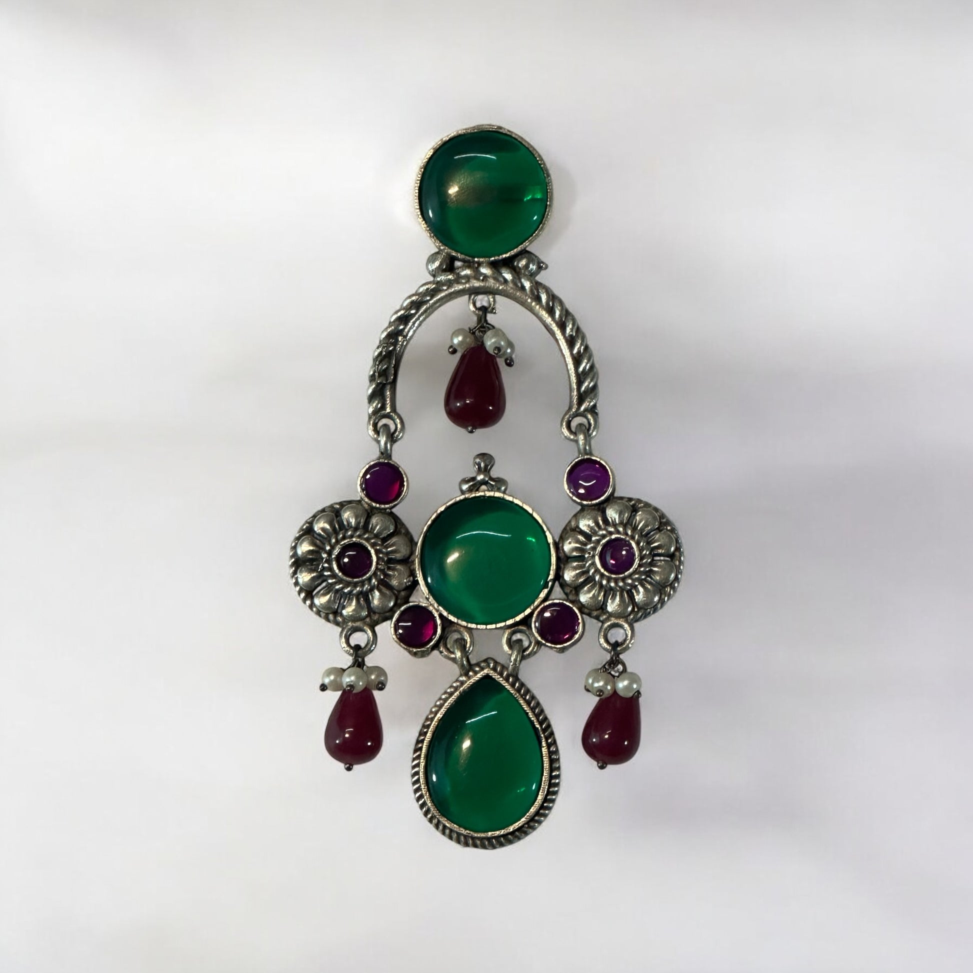 A pair of silver look alike designer earrings with red and green stones on a white  background.