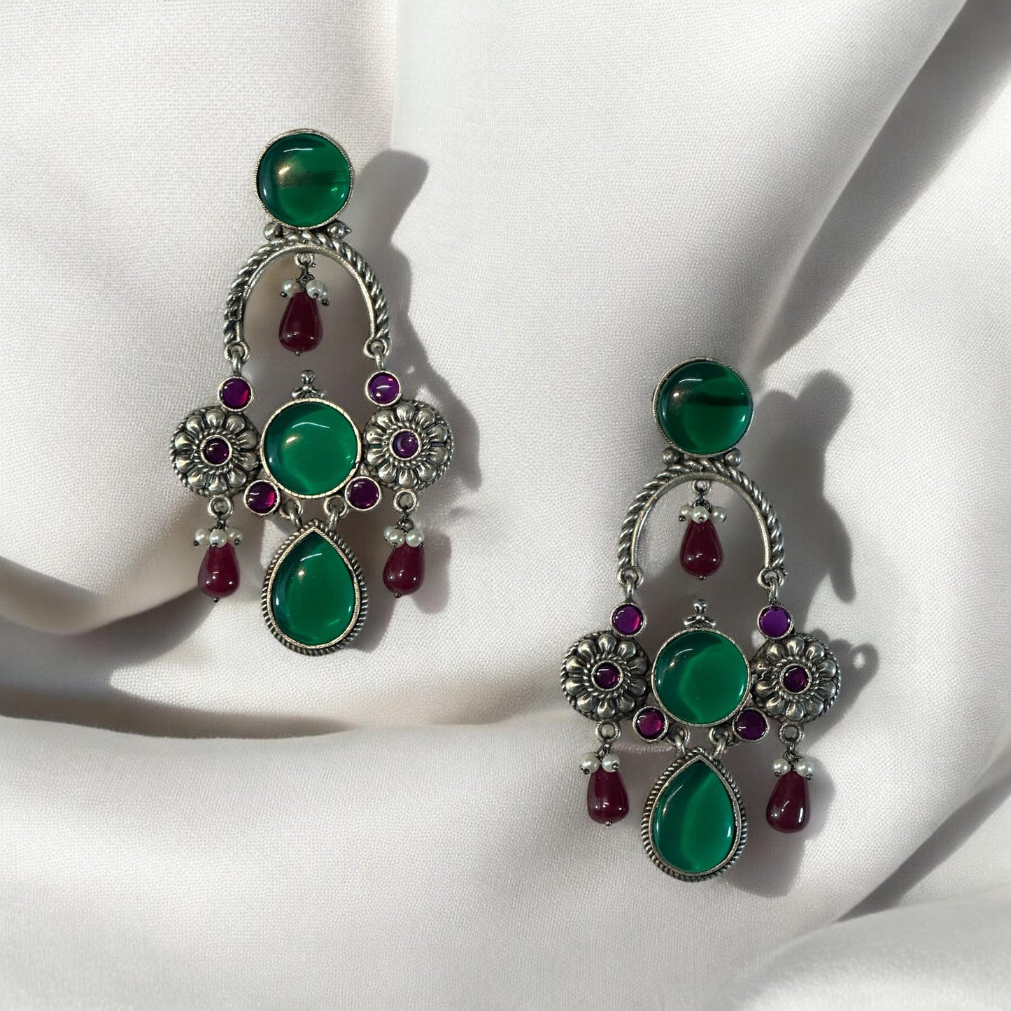 A pair of silver look alike designer earrings with red and green stones on a white fabric background.