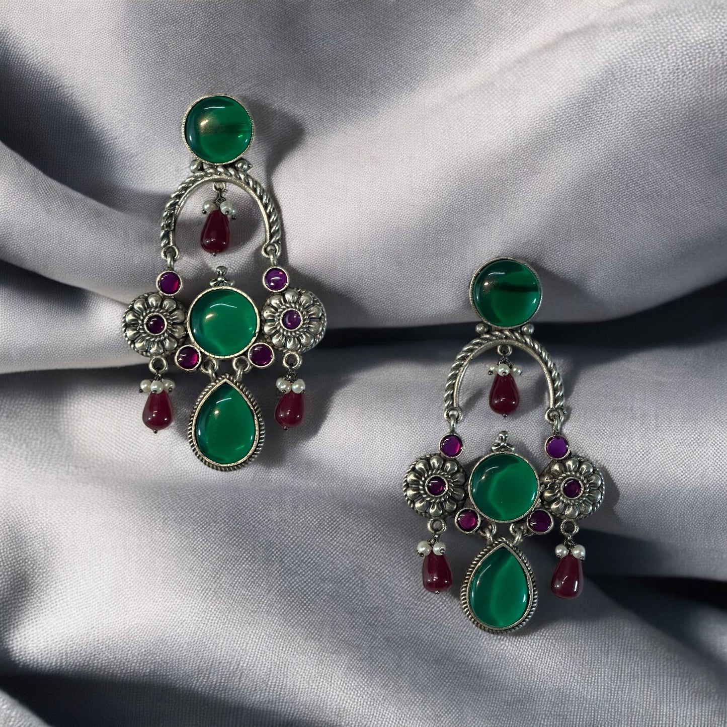 A pair of silver look alike designer earrings with red and green stones on a grey fabric background.