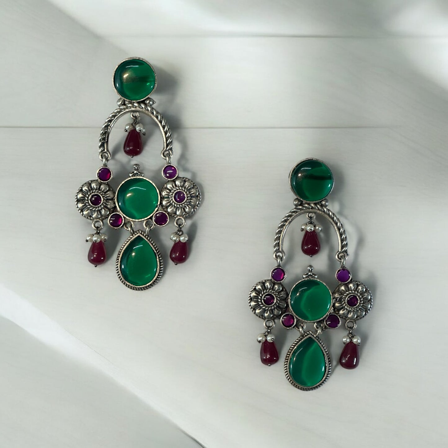 A pair of silver look alike designer earrings with red and green stones on a white fabric background.