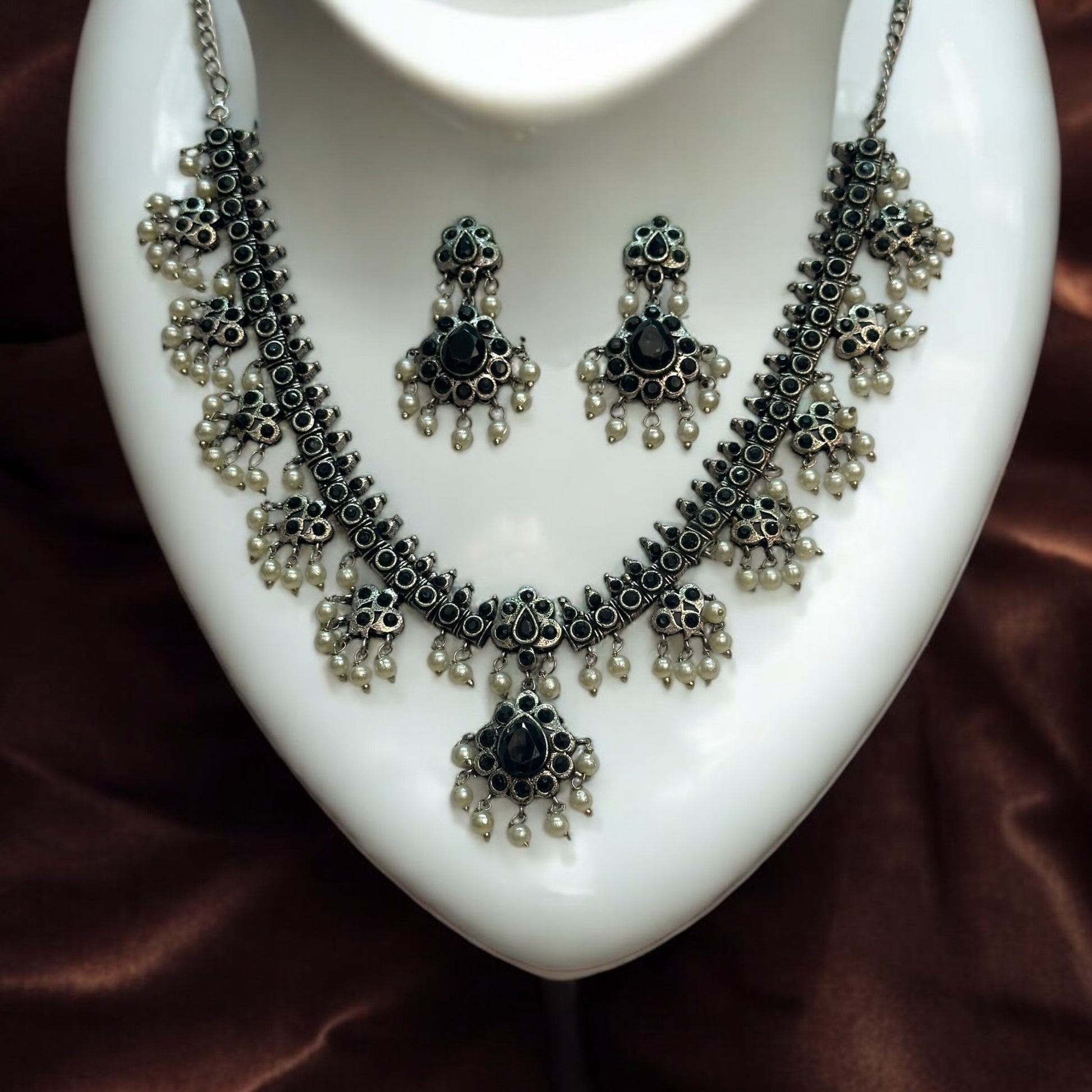 Silver Replica Necklace and Earrings Set with black accents, intricate detailing, and dangling pearl embellishments, displayed on a white jewellery display stand against a shiny dark brown fabric background.