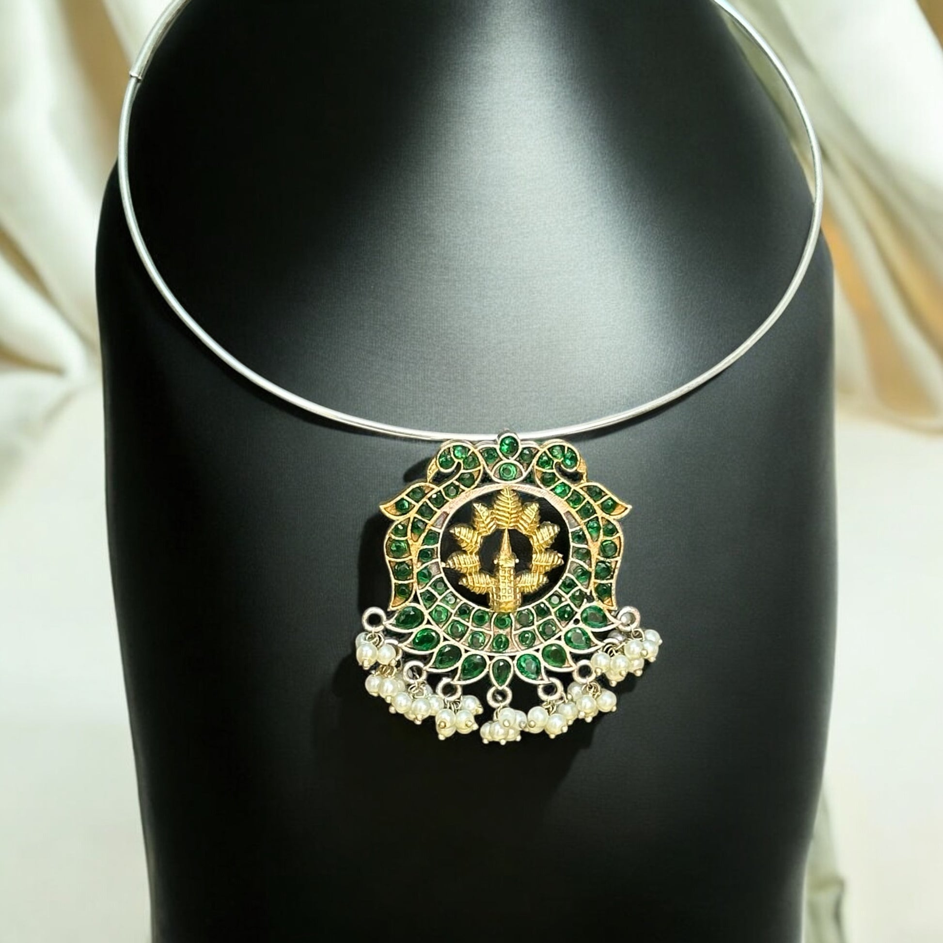 Silver replica Hasli necklace with a round green and gold statement pendant, adorned with pearl danglers, displayed on a black jewelry bust against a soft-lit background.