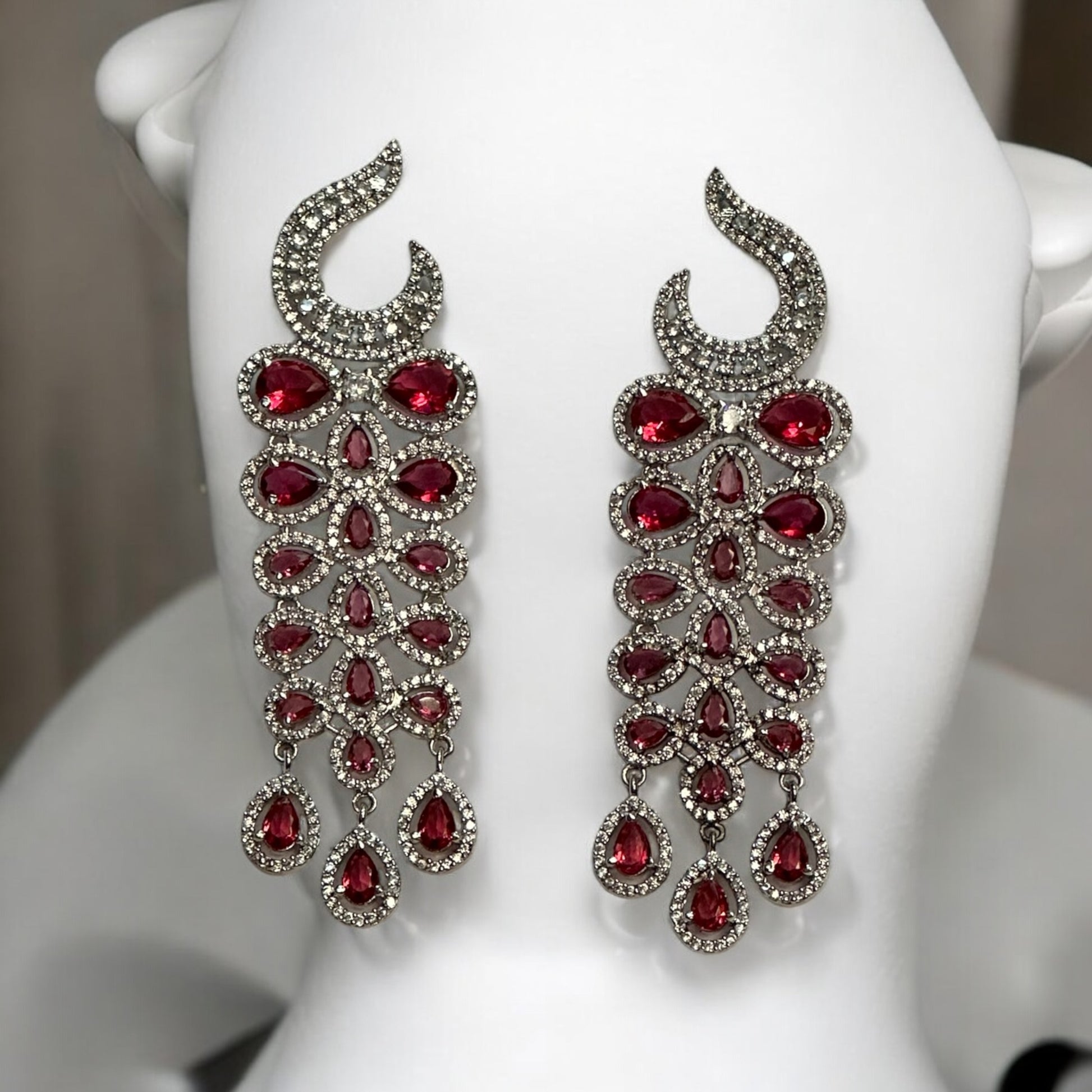 Statement Partywear American Diamond Earrings – stunning cascade earrings with sparkling American diamonds and ruby-red stones placed on a white coloured jewellery display stand. Perfect for weddings, cocktail parties, and festive events.