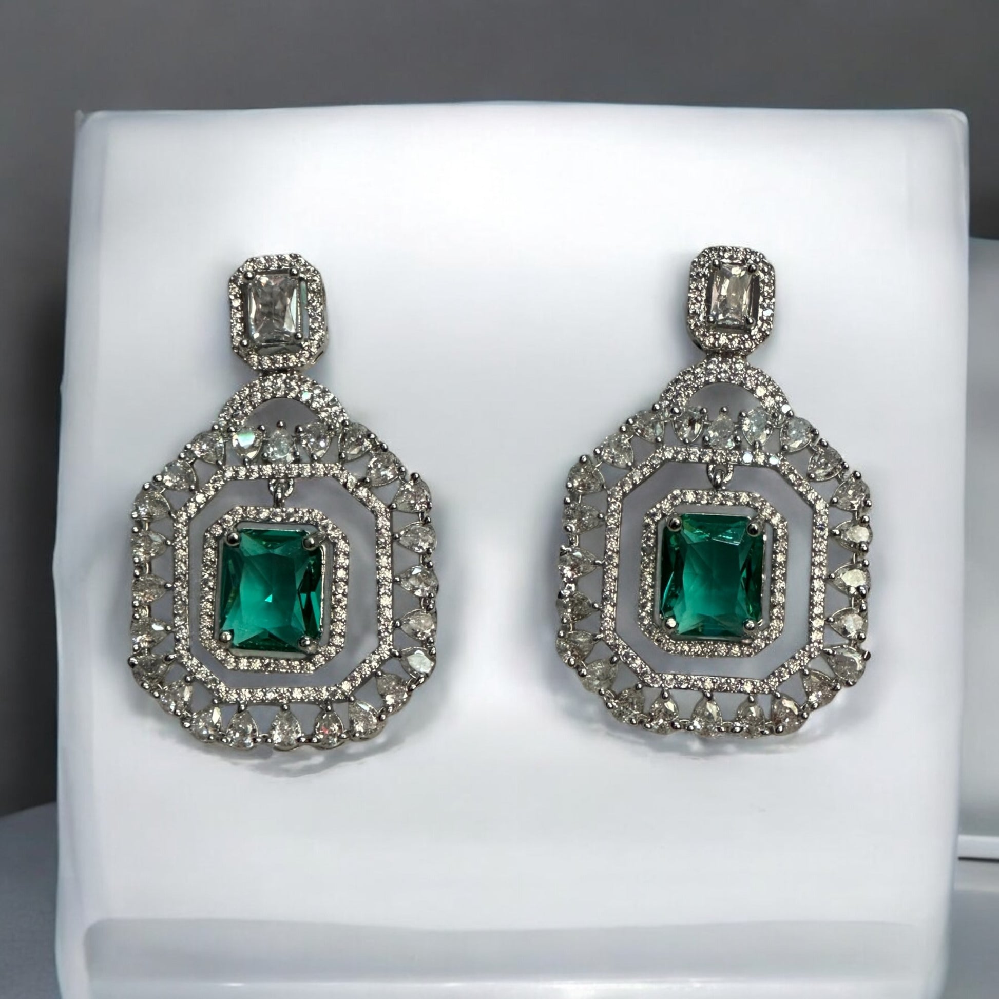 Beautiful Statement American Diamond Earrings – bold geometric earrings with sparkling American diamonds and emerald-green centerpiece stones placed on a jewellery display stand. Perfect for weddings and festive occasions.